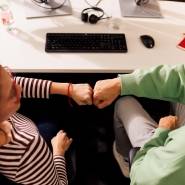 Kollege und Kollegin sitzen gemeinsam im Kaufland-Büro am Schreibtisch mit schwarzer Tastatur. Ihre zur Faust geballten Hände berühren sich zum Gruß.