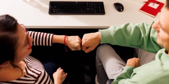 Kollege und Kollegin sitzen gemeinsam im Kaufland-Büro am Schreibtisch mit schwarzer Tastatur. Ihre zur Faust geballten Hände berühren sich zum Gruß.