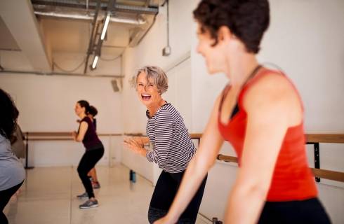Zwei Frauen nehmen an einem Fitnesskurs teil und lachen dabei. Im Hintergrund sieht man weitere Teilnehmer.