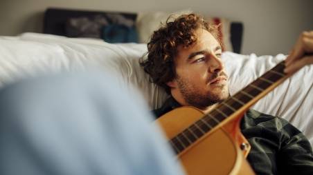 Mann sitzt auf dem Boden vor einem Bett und spielt Gitarre.